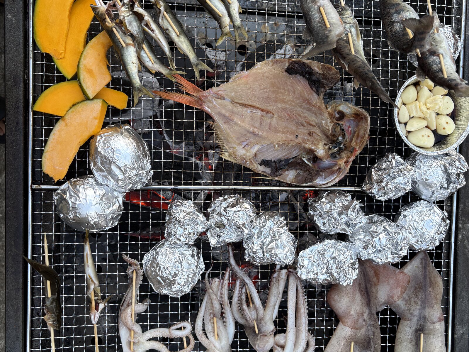 炭火の網一面に並んだ川魚・イカ・ホイル焼き・かぼちゃ